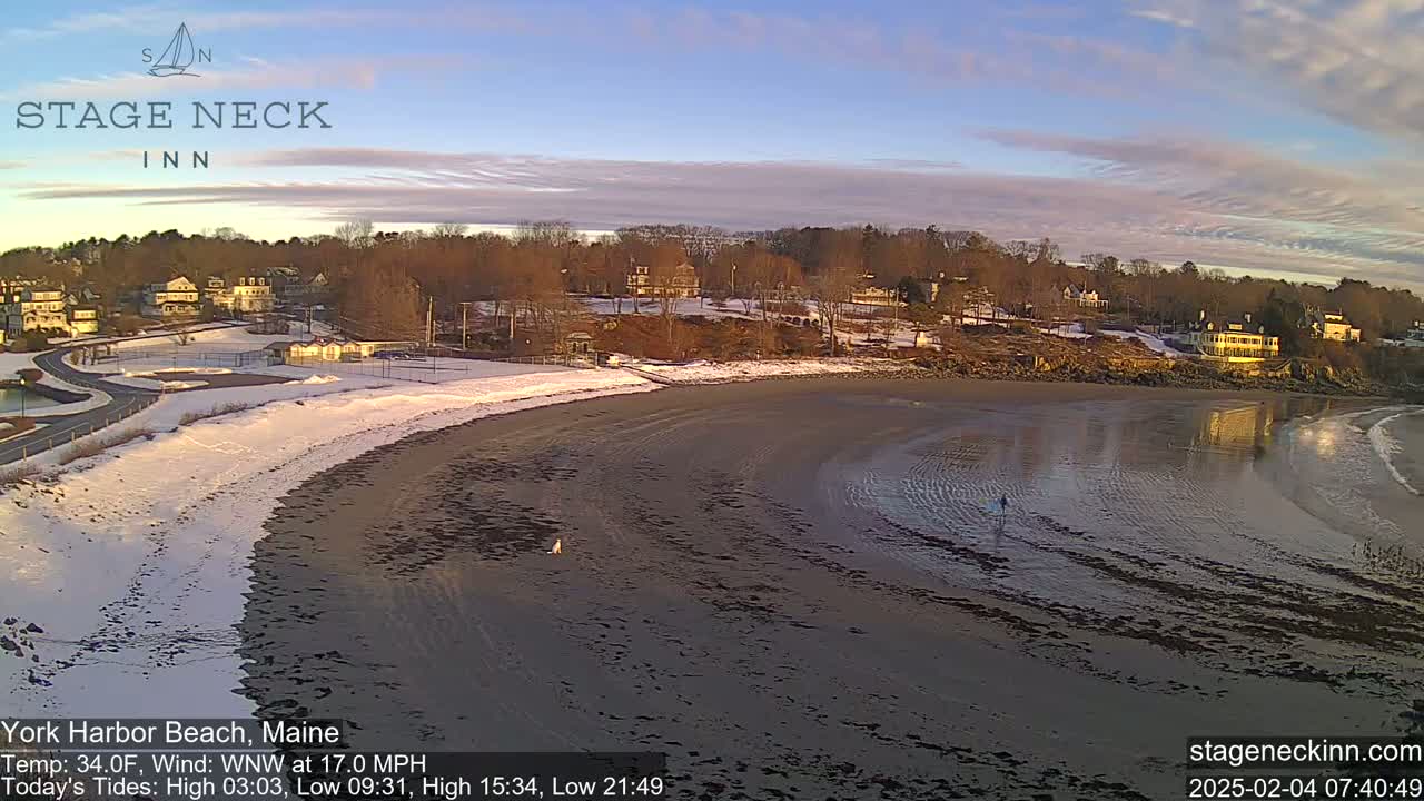 A sandy beach, partially covered in snow, curves alongside a residential area under a partly cloudy, sunrise sky.