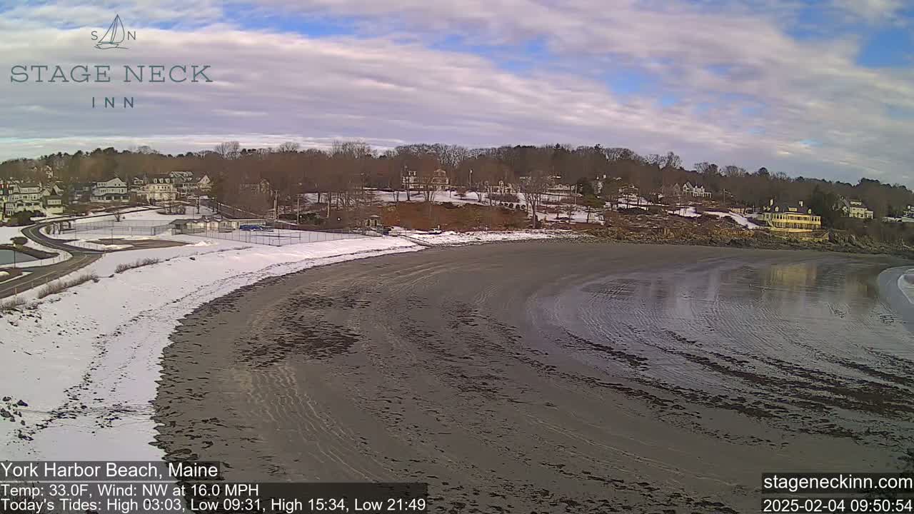 A partially snow-covered beach with a receding tide is seen under a partly cloudy sky, with buildings and trees visible in the background.