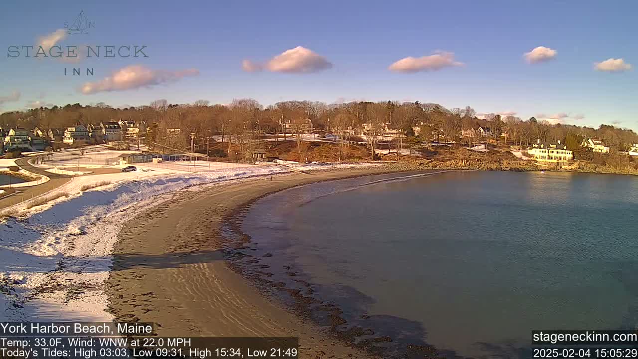 A sandy beach partially covered in snow curves into a calm body of water under a mostly clear sky with a few scattered clouds.