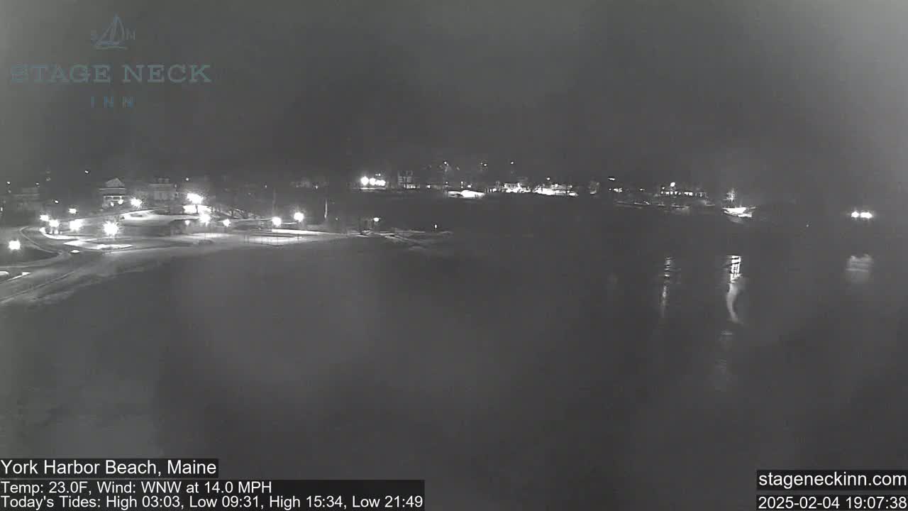 A nighttime view of a snow-covered coastal area with faint lights from buildings and a calm body of water reflecting the lights.