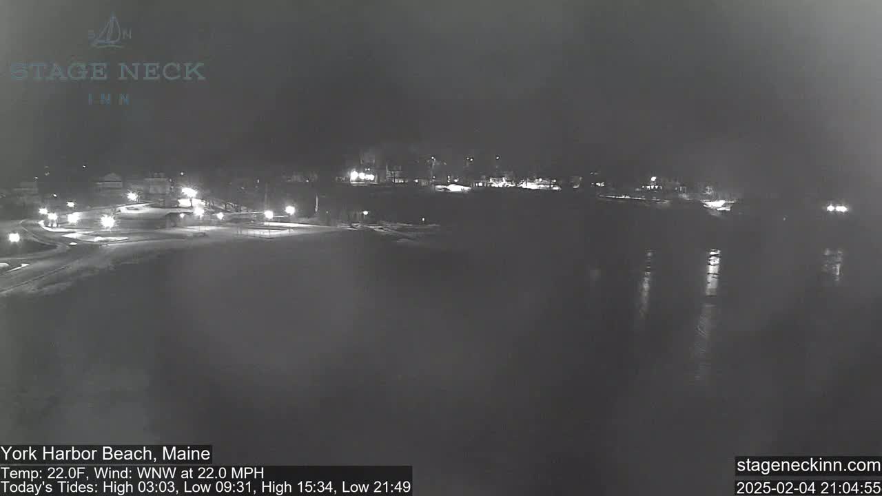A nighttime view of a snow-covered beach and waterfront area with faint lights reflecting on dark water under a dark sky.