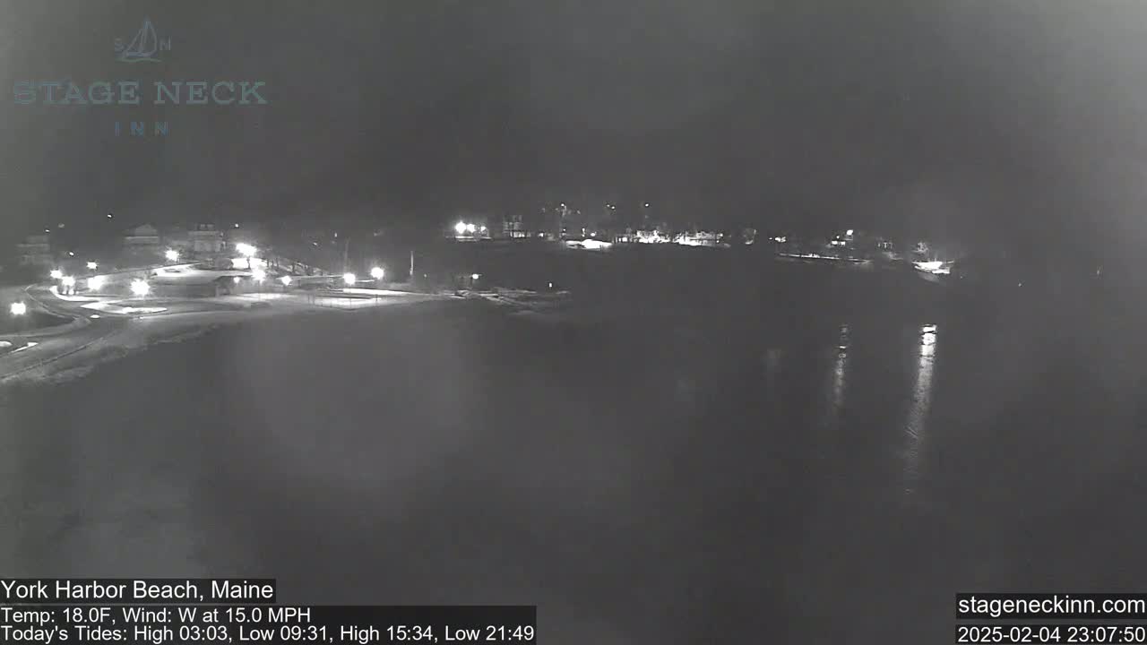 A nighttime, low-light view of a snow-covered beach and waterfront area with scattered lights, under a dark sky with a 15 mph wind.