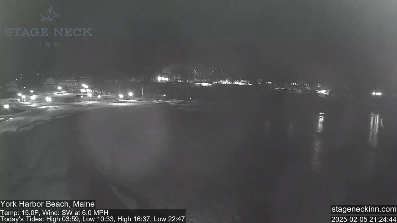 A nighttime view of a snow-covered beach and calm water, with dimly lit buildings and streets in the background under a dark sky.