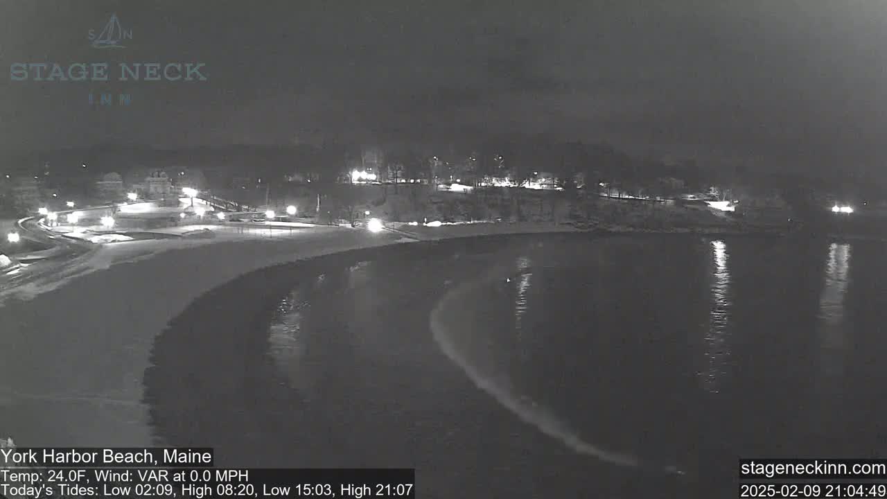 A snow-covered beach at night is partially lit by streetlights, with calm dark water lapping at the shore.
