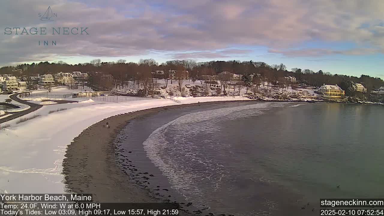 A snow-covered beach with a calm ocean and houses on a hillside under a partly cloudy sky.