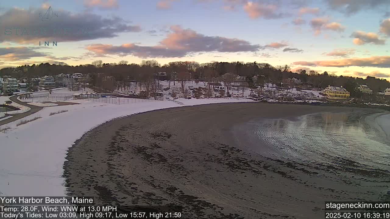 A partially snow-covered beach and residential area are shown at sunset under a partly cloudy sky.