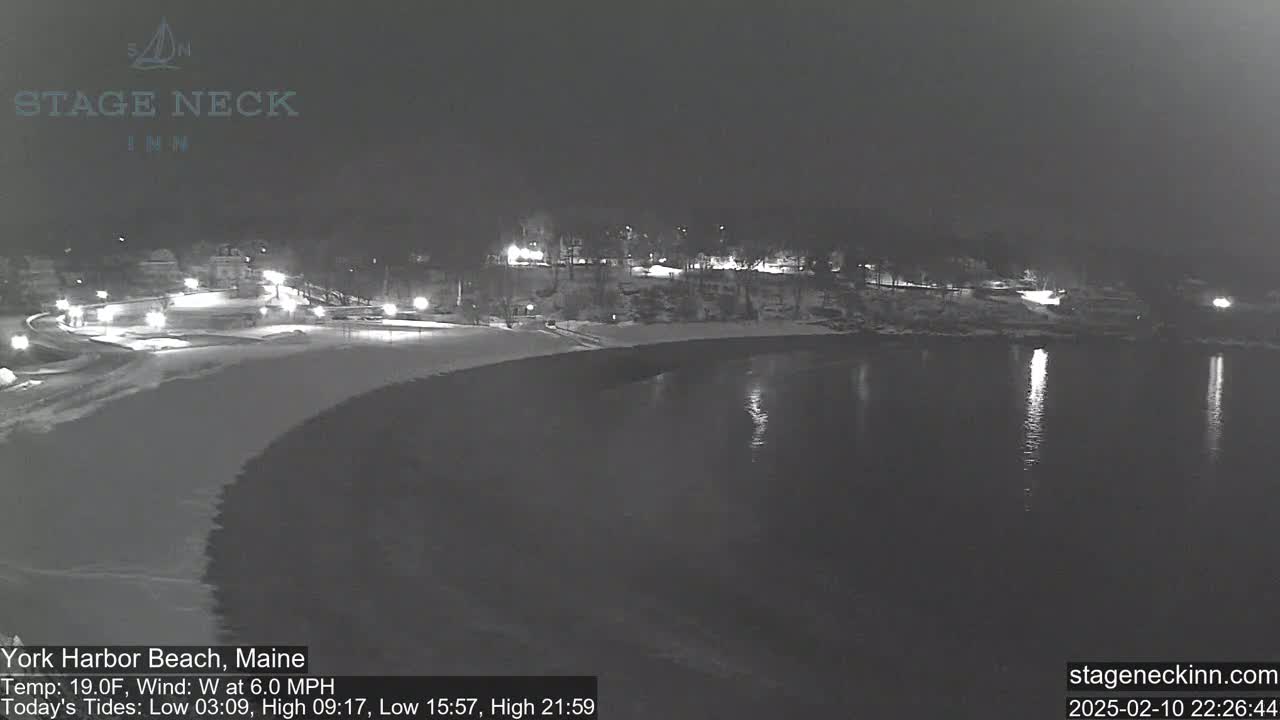 A snow-covered beach at night is illuminated by lights from buildings across a calm body of water under a dark sky.