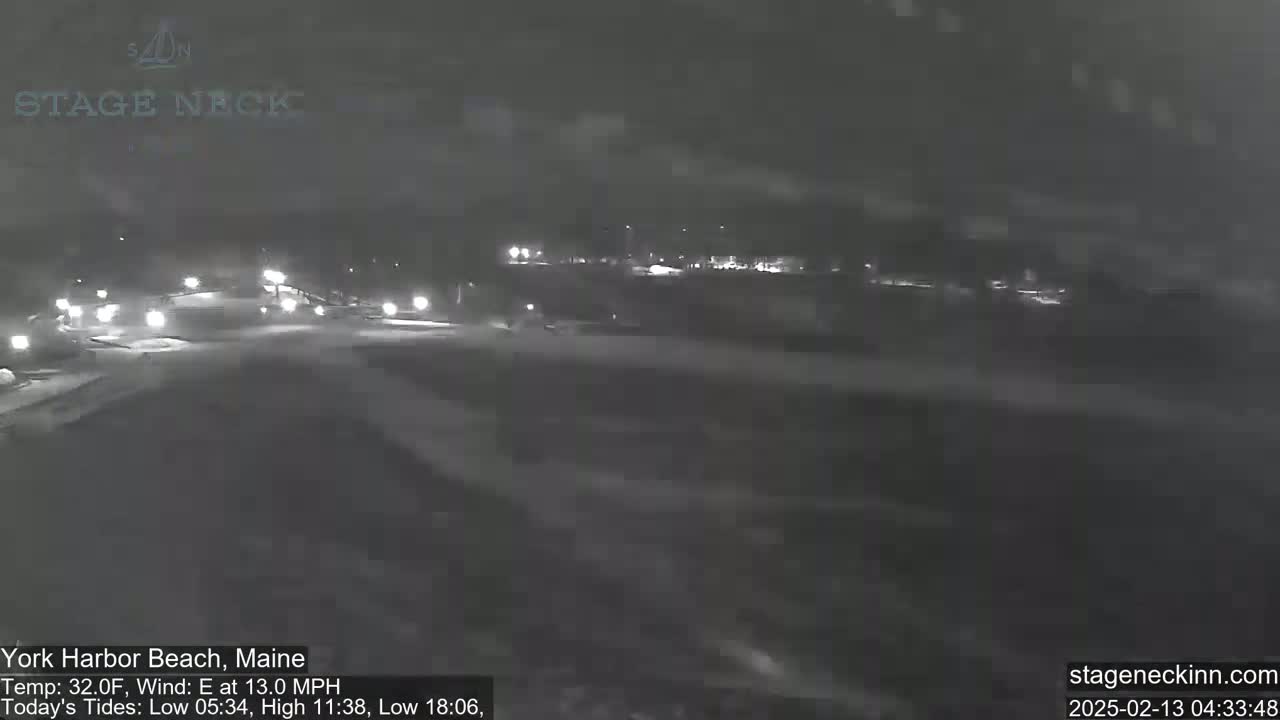 A nighttime, snowy scene shows a road with a few cars driving through a dimly lit area.