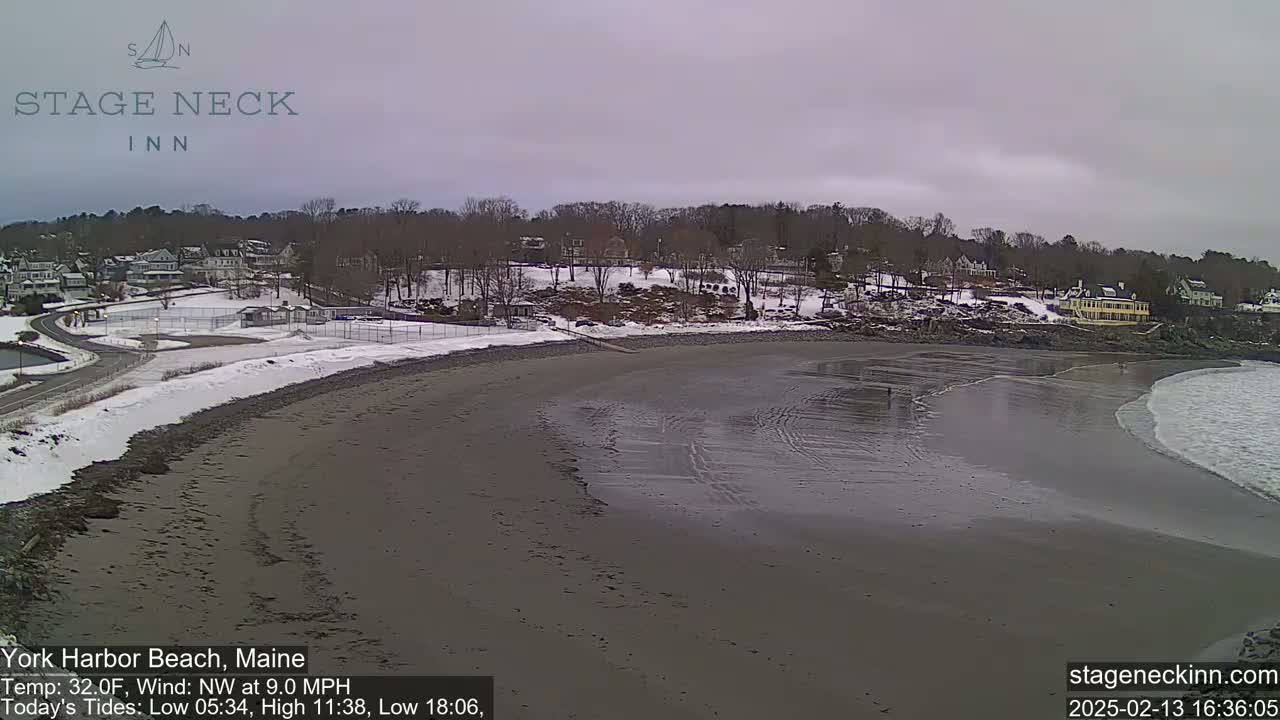 A sandy beach with some snow on the shore and partially frozen water, overlooks a snow-covered town under an overcast sky.