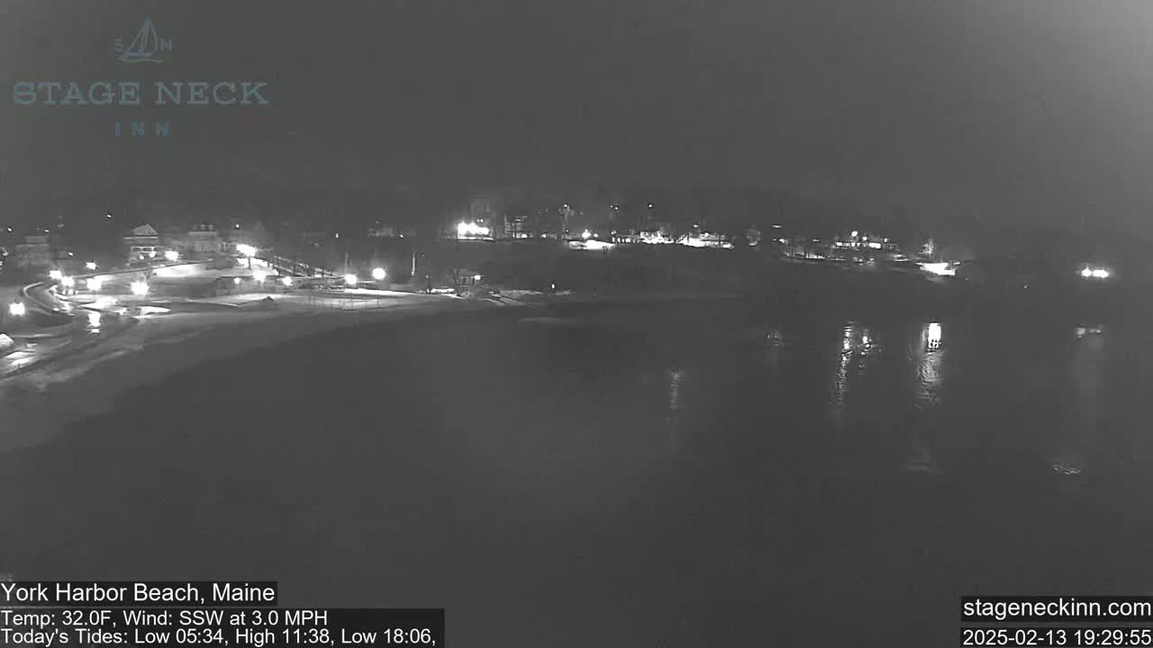 A nighttime view of a snow-covered beach and calm water with dimly lit buildings in the background.