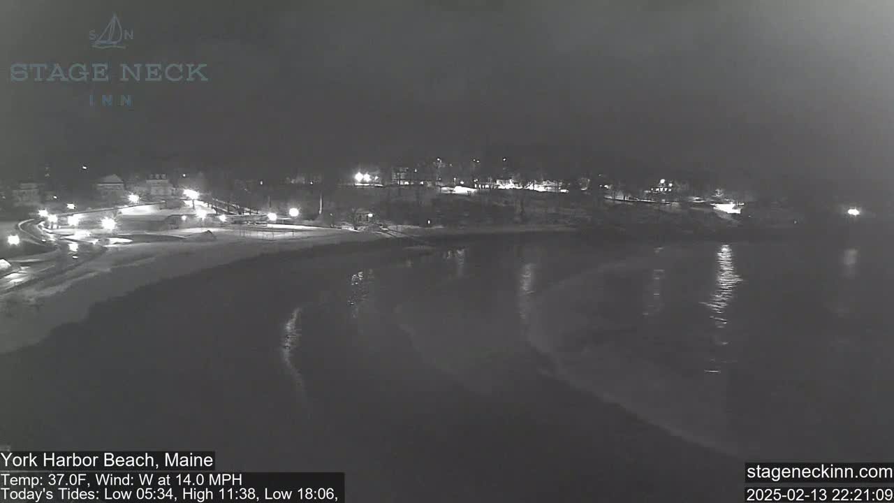 A nighttime view of a snow-covered beach and calm water, with lights visible along a shoreline and in the distance.