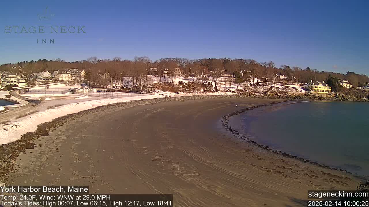 A sandy beach curves along a calm bay, with a snow-dusted shoreline and houses nestled on a hill in the background under a clear blue sky.
