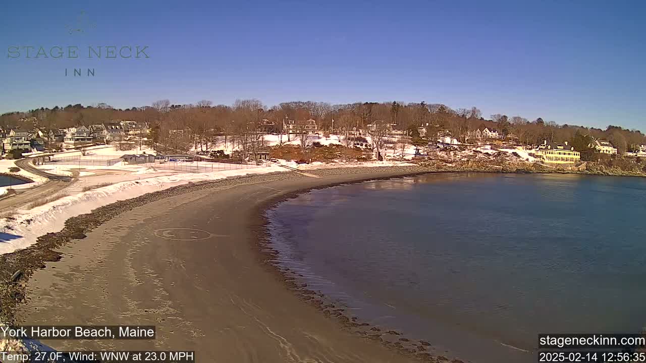 A sandy beach curves along a calm body of water under a clear blue sky, with a snowy shoreline and buildings in the background.