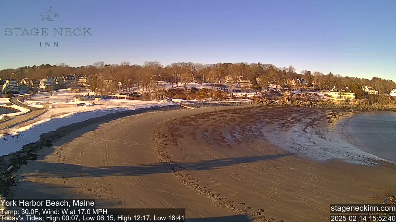 A sandy beach with receding water and a partially snow-covered shoreline curves around a residential area under a clear, sunny sky.