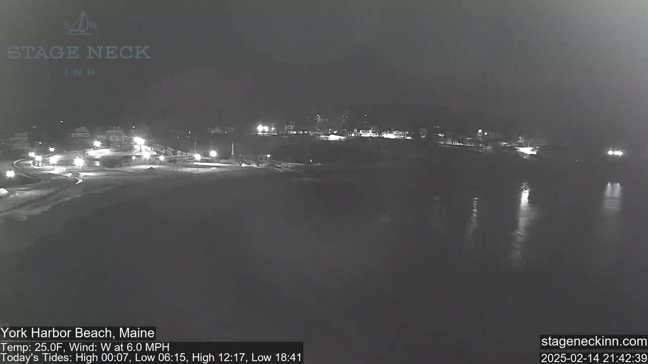 A nighttime view of a snow-covered beach and shoreline with dimly lit buildings and a calm body of water reflecting the lights.
