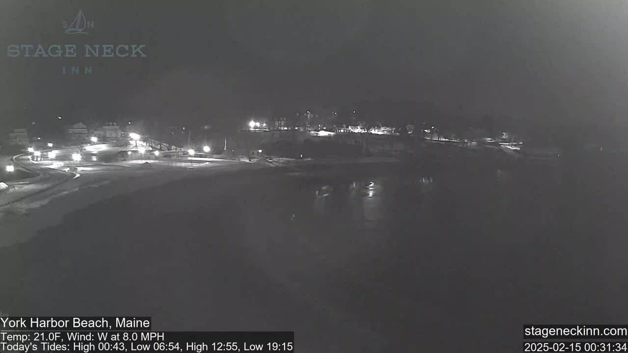 A nighttime view of a snow-covered beach and waterfront area with dimly lit buildings and a calm body of water.