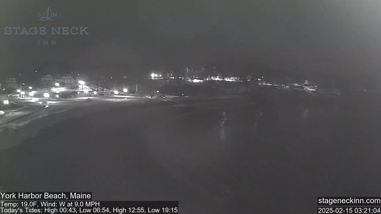A nighttime, snow-covered coastal scene with sparsely lit buildings and a dark body of water.
