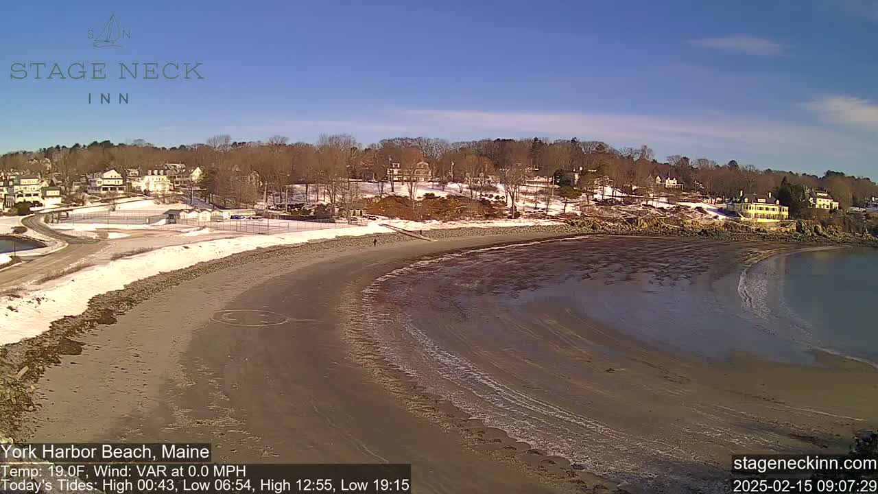 A sandy beach curves gently into calm water under a clear blue sky, with snow visible on the land and some buildings in the distance.