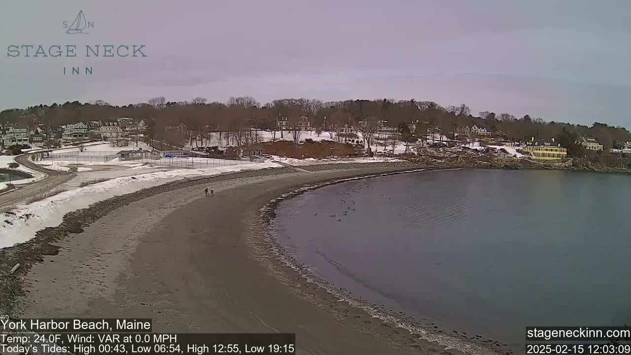 A mostly sandy beach curves gently into a calm body of water under an overcast sky, with a snow-dusted shoreline and buildings visible in the background.