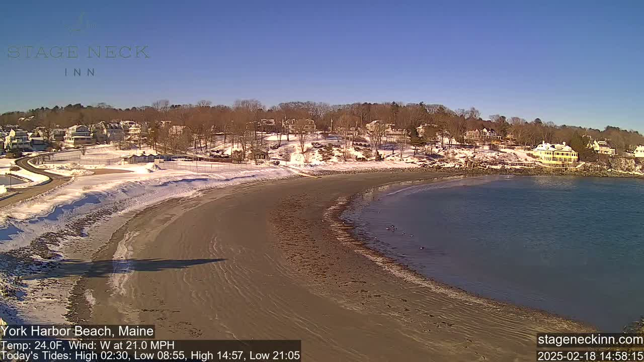 A sandy beach curves into a calm bay under a clear blue sky, with snow on the ground and buildings lining the shore.