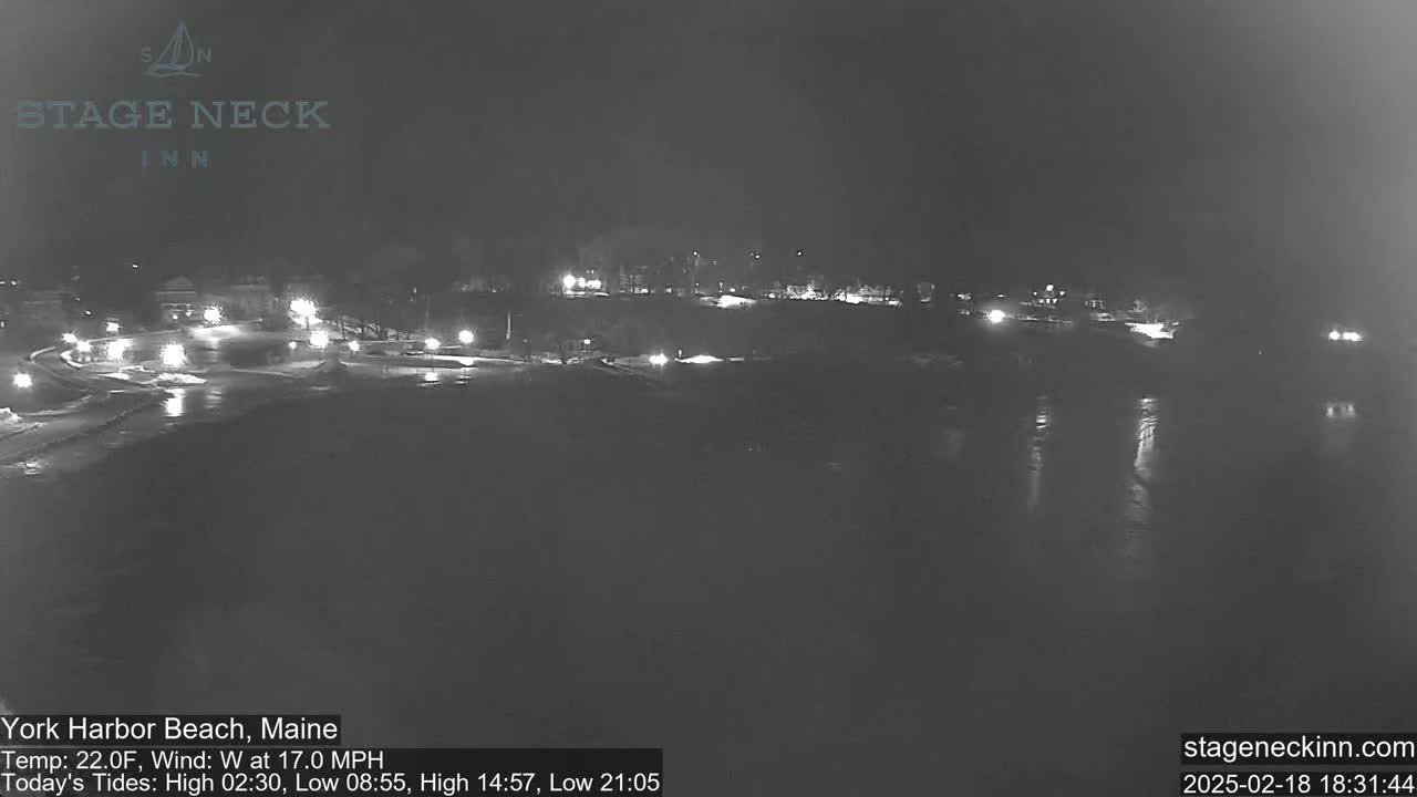 A nighttime view of a snow-covered beach and road with some distant lights and a calm body of water reflecting the lights.
