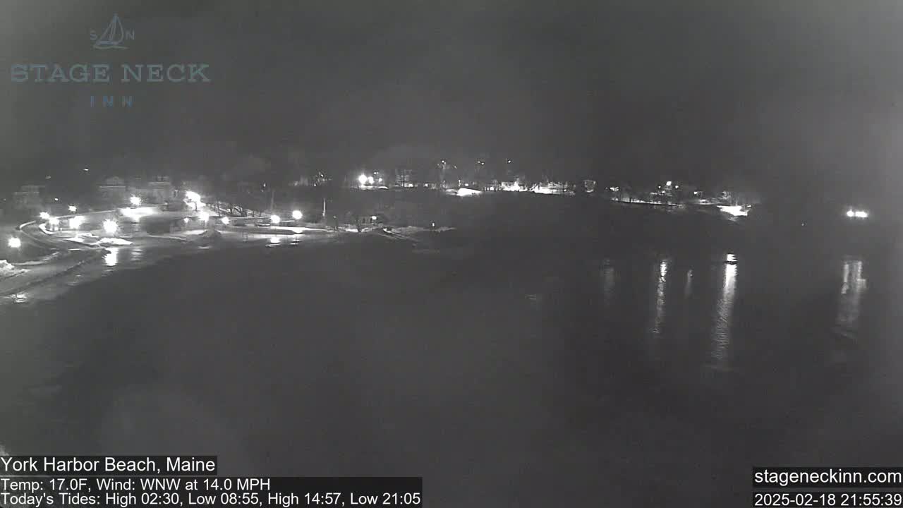 A nighttime view of a snow-covered beach and calm water reflecting lights from a nearby town under a dark sky.