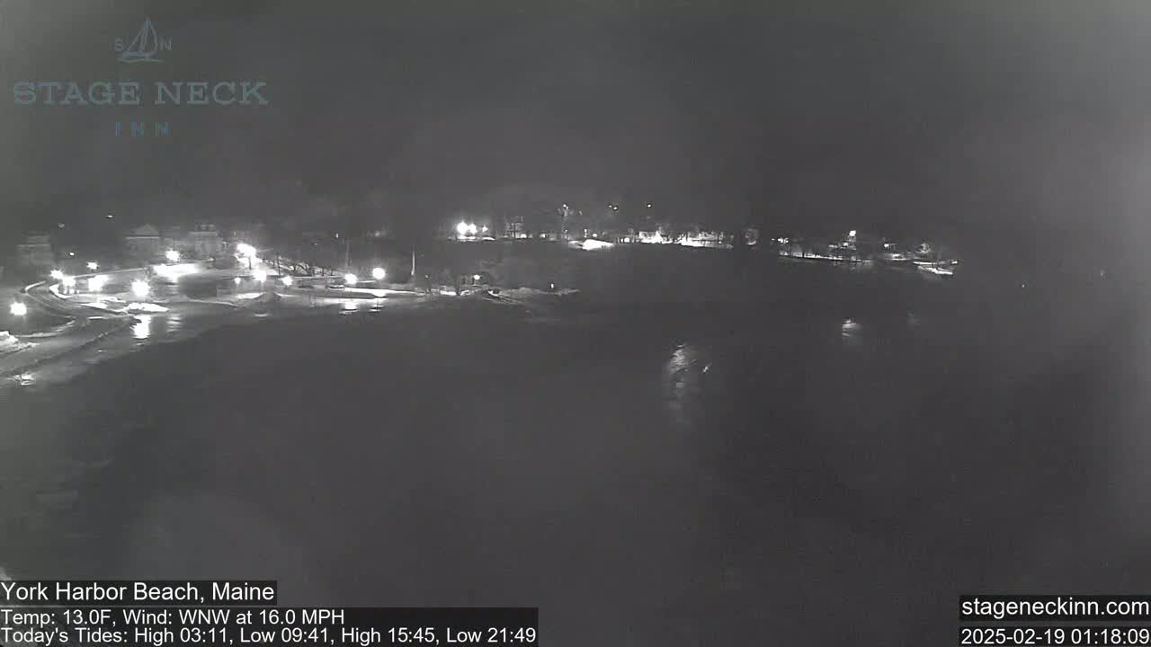 A nighttime view of a snow-covered beach and town, illuminated by scattered lights, under a dark, cloudy sky.