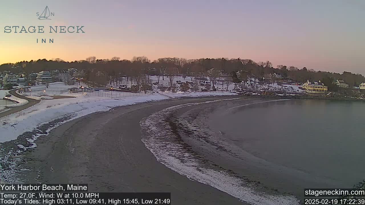 A snow-dusted beach curves into a calm bay under a pale pink and purple twilight sky.