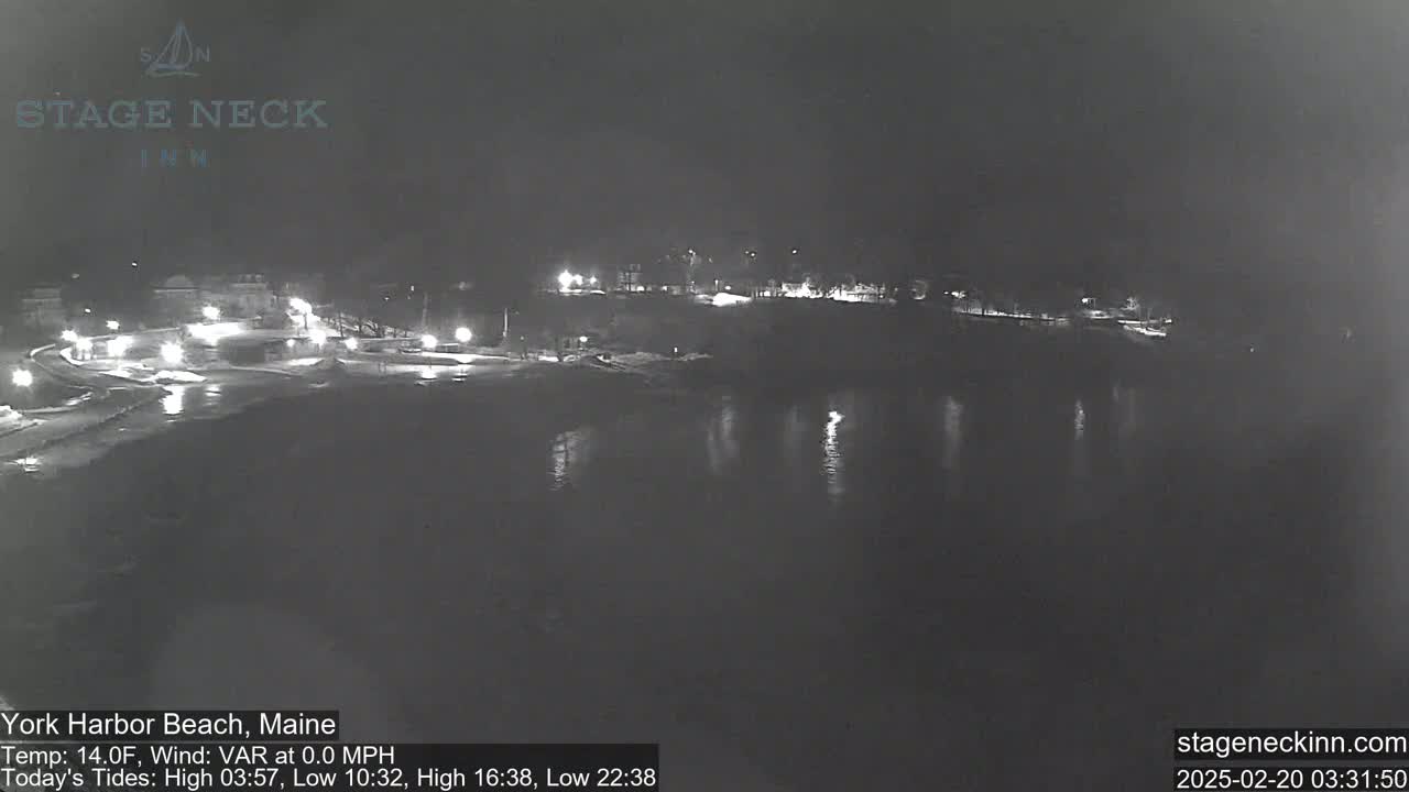 A nighttime, low-light view of a dark, calm body of water reflecting lights from a shoreline with buildings and trees.