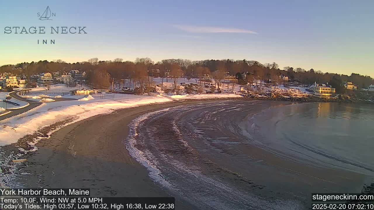A snow-covered beach curves into a calm bay under a clear, sunrise sky.