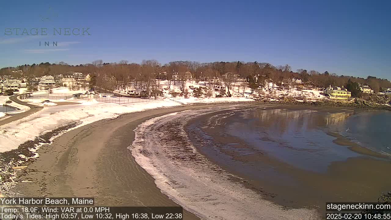 A sandy beach curves along a calm, dark-brown ocean under a clear blue sky, with a snow-dusted shoreline and houses visible in the background.