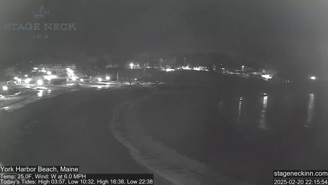 A nighttime view of a snow-covered beach and calm water, with lights illuminating a roadway and buildings in the distance.