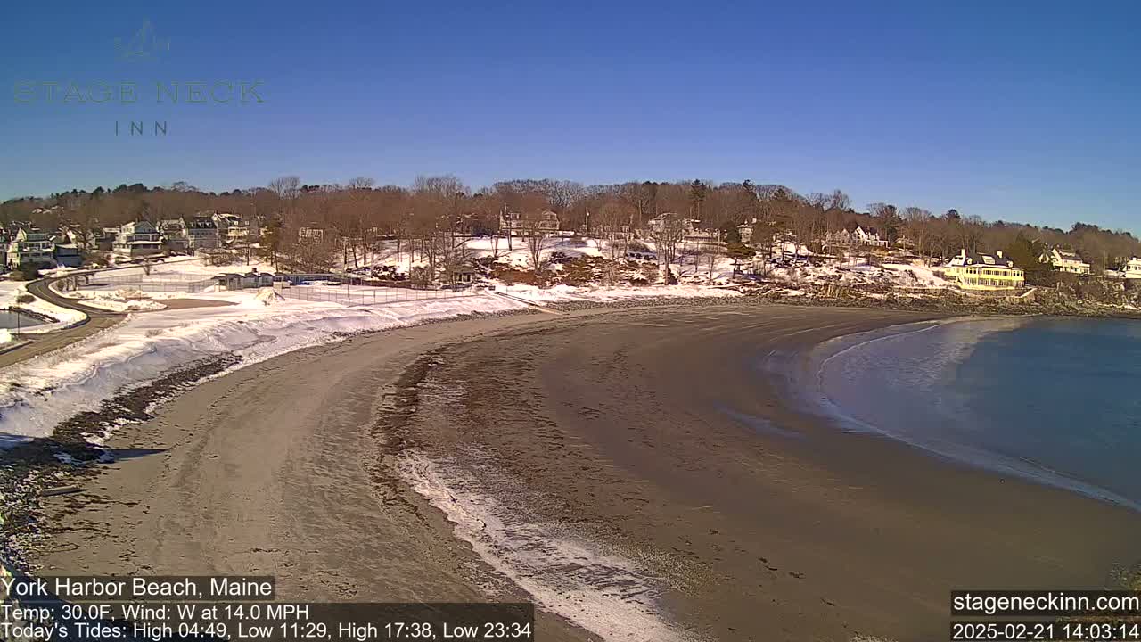 A sandy beach curves along a calm ocean under a clear blue sky, with snow on the ground and nearby buildings partially visible behind a snow-covered hill.