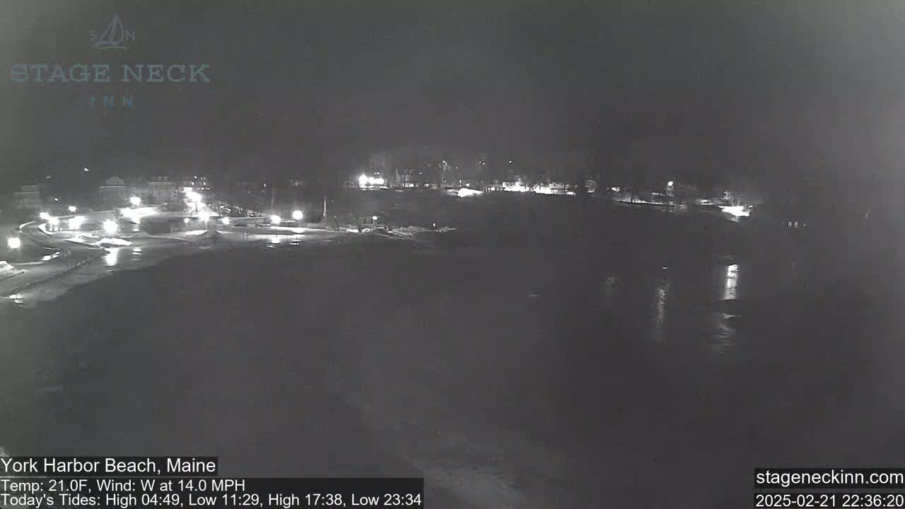 A nighttime view of a snow-covered coastal area with dimly lit buildings and a dark body of water reflecting lights, under a cloudy sky.