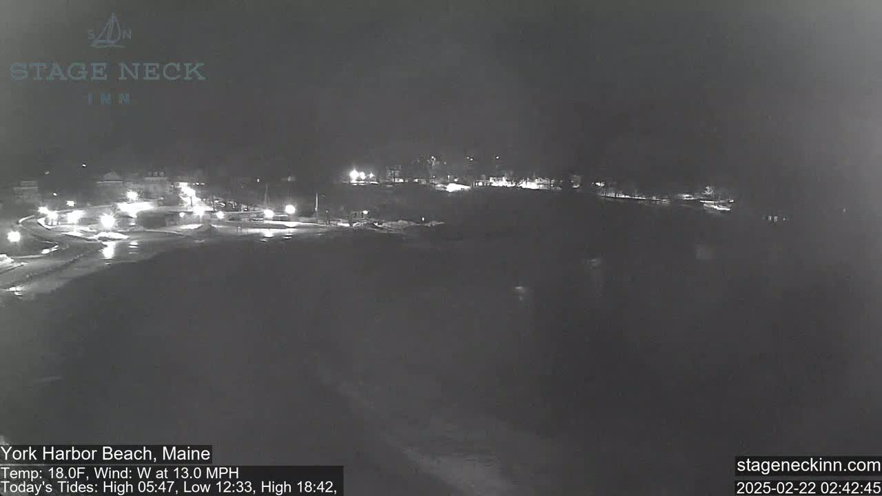 A nighttime view of a snow-covered parking lot and roadway with some distant lights, under a dark sky with a west wind.