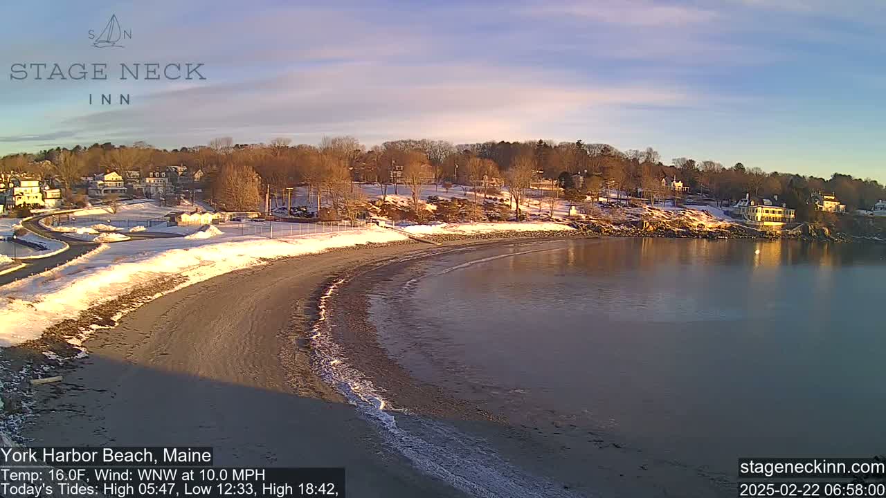 A partially snow-covered beach curves into a calm body of water under a clear, twilight sky.