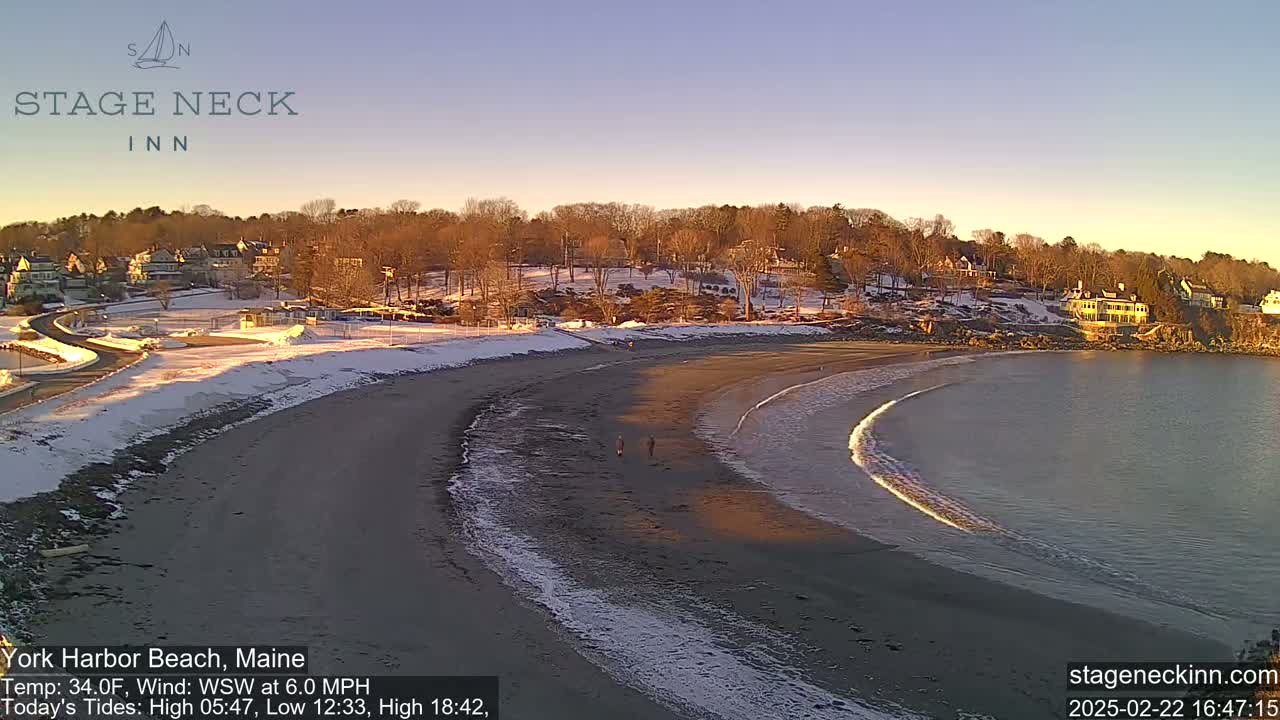 A sandy beach curves into a calm bay under a clear, twilight sky, with snow dusting the shoreline and nearby buildings.