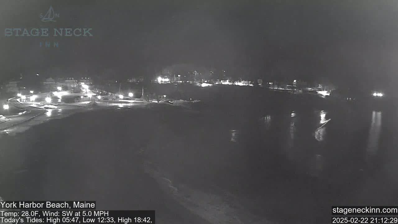 A nighttime, low-light view of a coastal area shows a dark body of water reflecting sparse lights from buildings and vehicles in the distance under a dark, overcast sky.