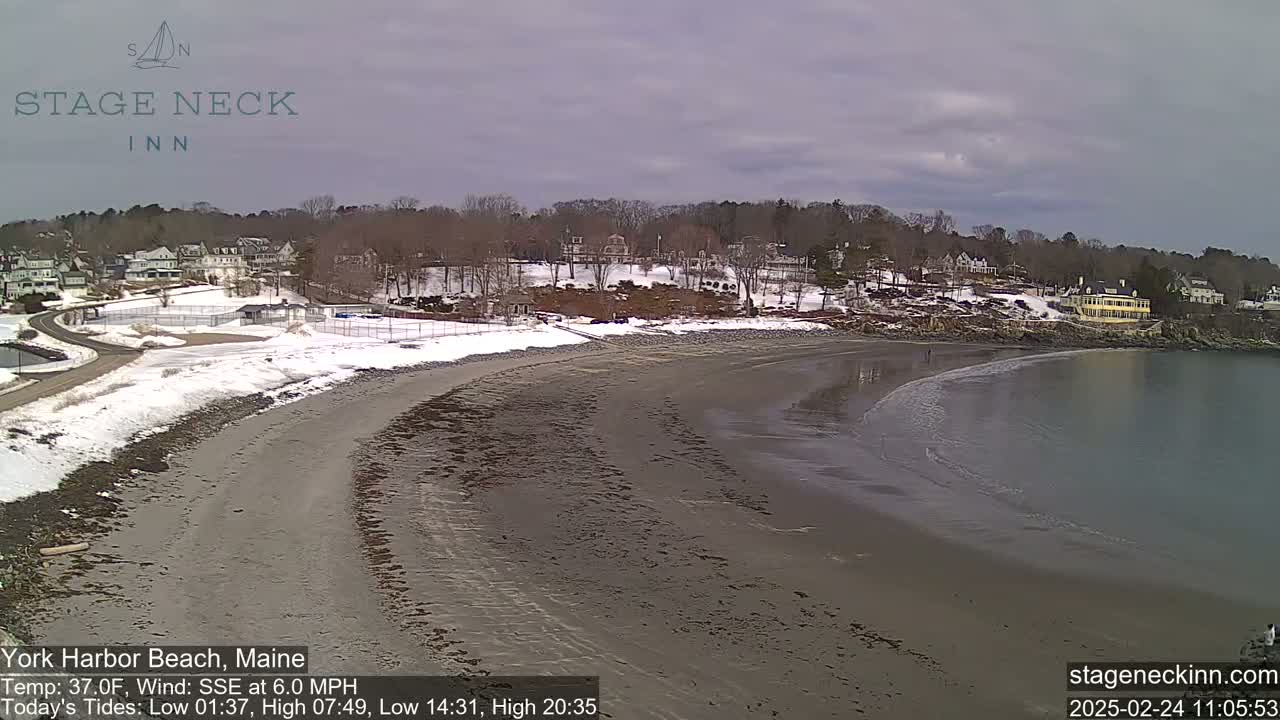 A partly cloudy day shows a sandy beach curving into a calm bay, with a snow-dusted shoreline and buildings in the background.