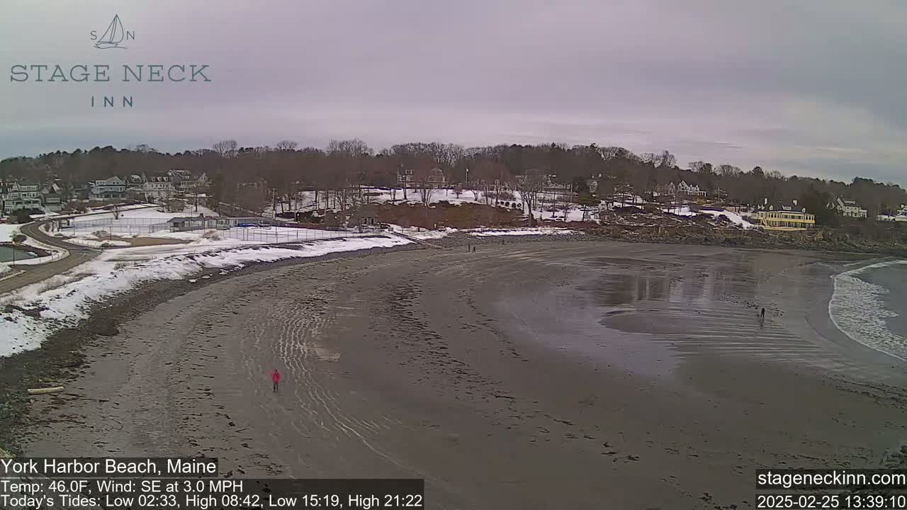 A wide sandy beach, partially covered in snow, stretches along a coastline under an overcast sky, with a few people walking along the shore and houses visible in the distance.