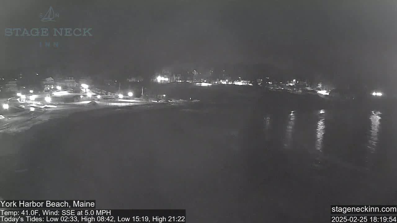 A nighttime view of a snow-covered beach and calm water, with some lights visible in the distance.
