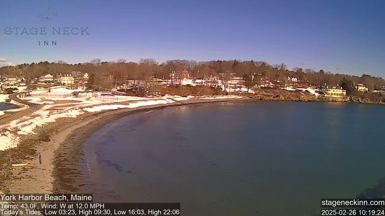 A partly snowy beach curves along a calm bay, with houses and trees visible on the hillside beyond.
