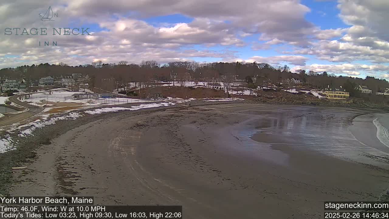 A mostly cloudy day shows a sandy beach with a receding tide,  buildings and trees are visible in the background, with some snow on the ground near the buildings.