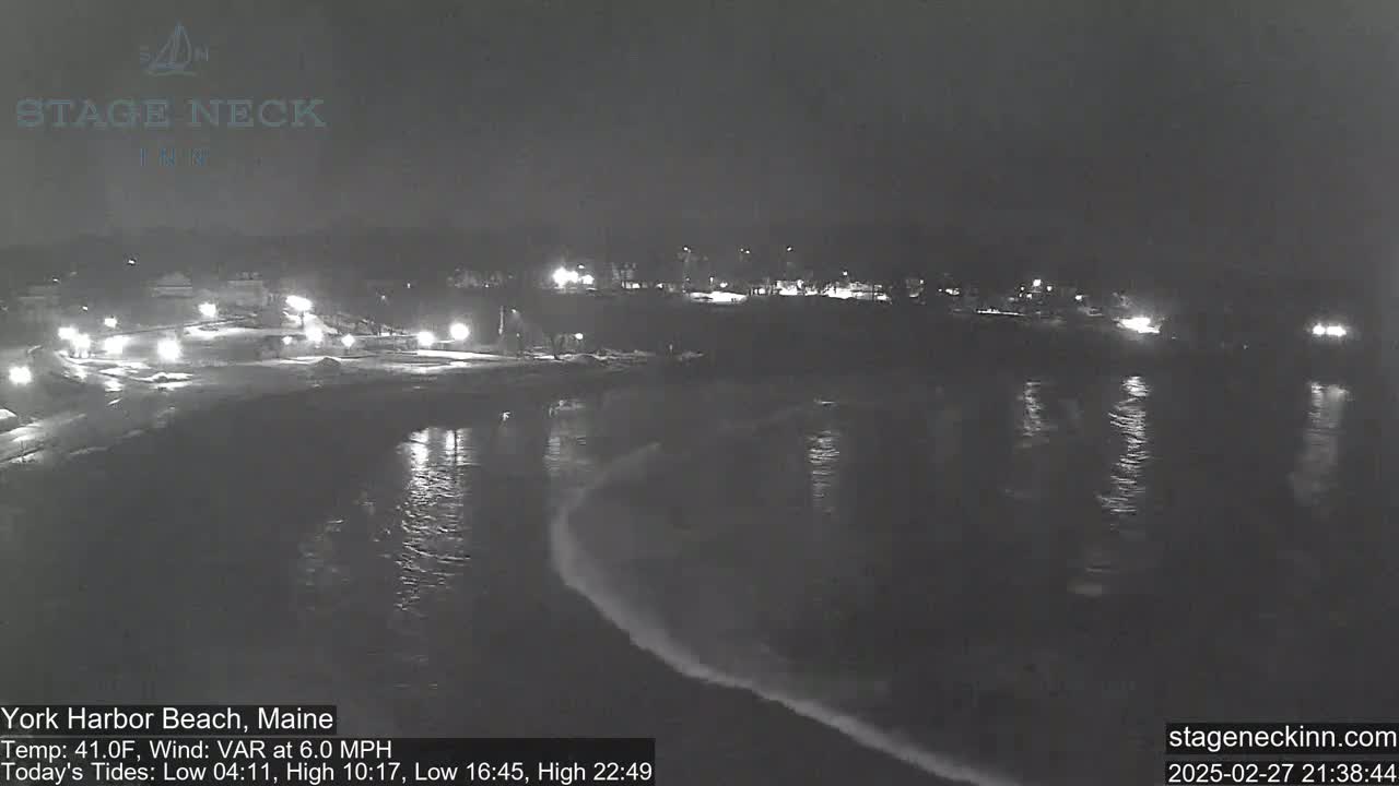 A nighttime view of a dark, wavy ocean shore with a snow-covered roadway and dimly lit buildings in the background under a dark sky.