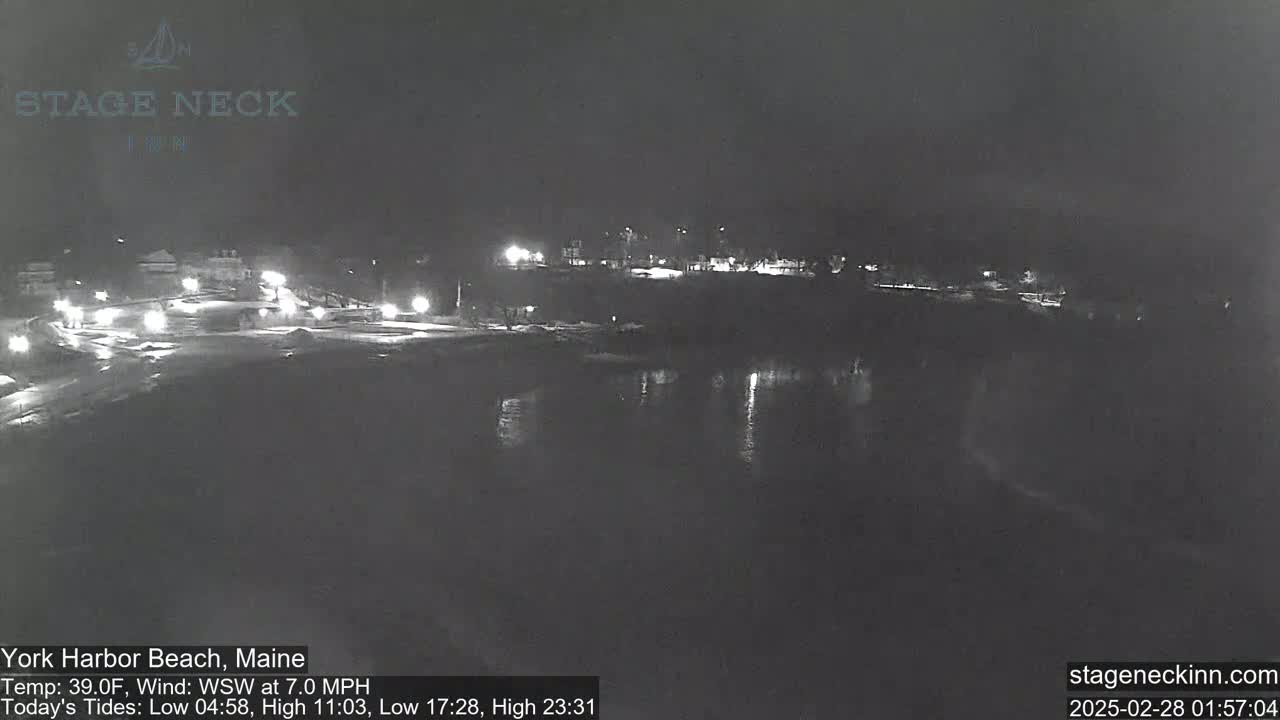 A nighttime, snow-covered coastal scene shows a body of dark water reflecting sparse lights from a distant roadway and buildings under a dark sky.