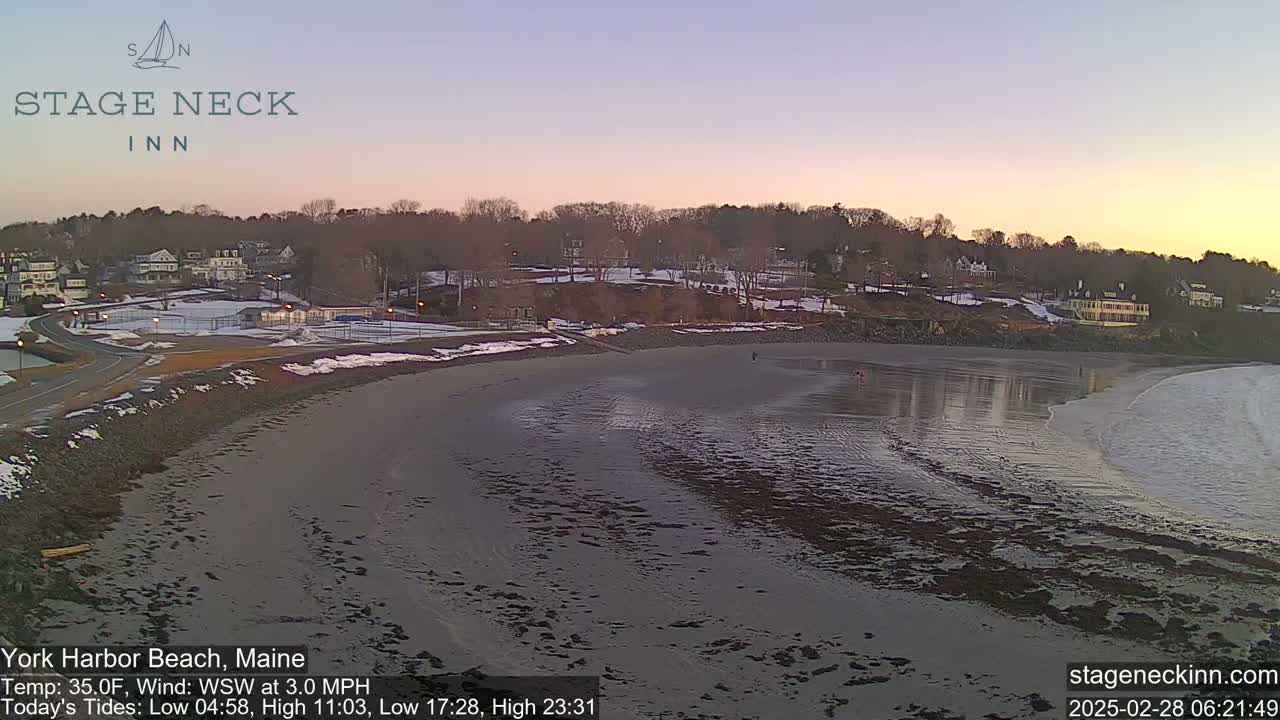 A mostly sandy beach with a calm ocean and small waves at sunset, overlooking a snow-dusted town with houses and trees.