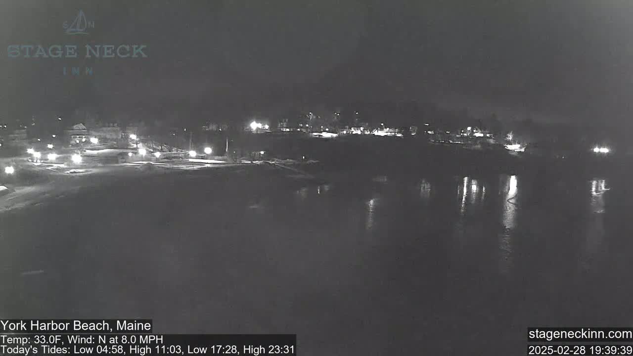 A nighttime view of a dark, calm body of water reflecting a few lights from buildings across the shore, under a clear sky.