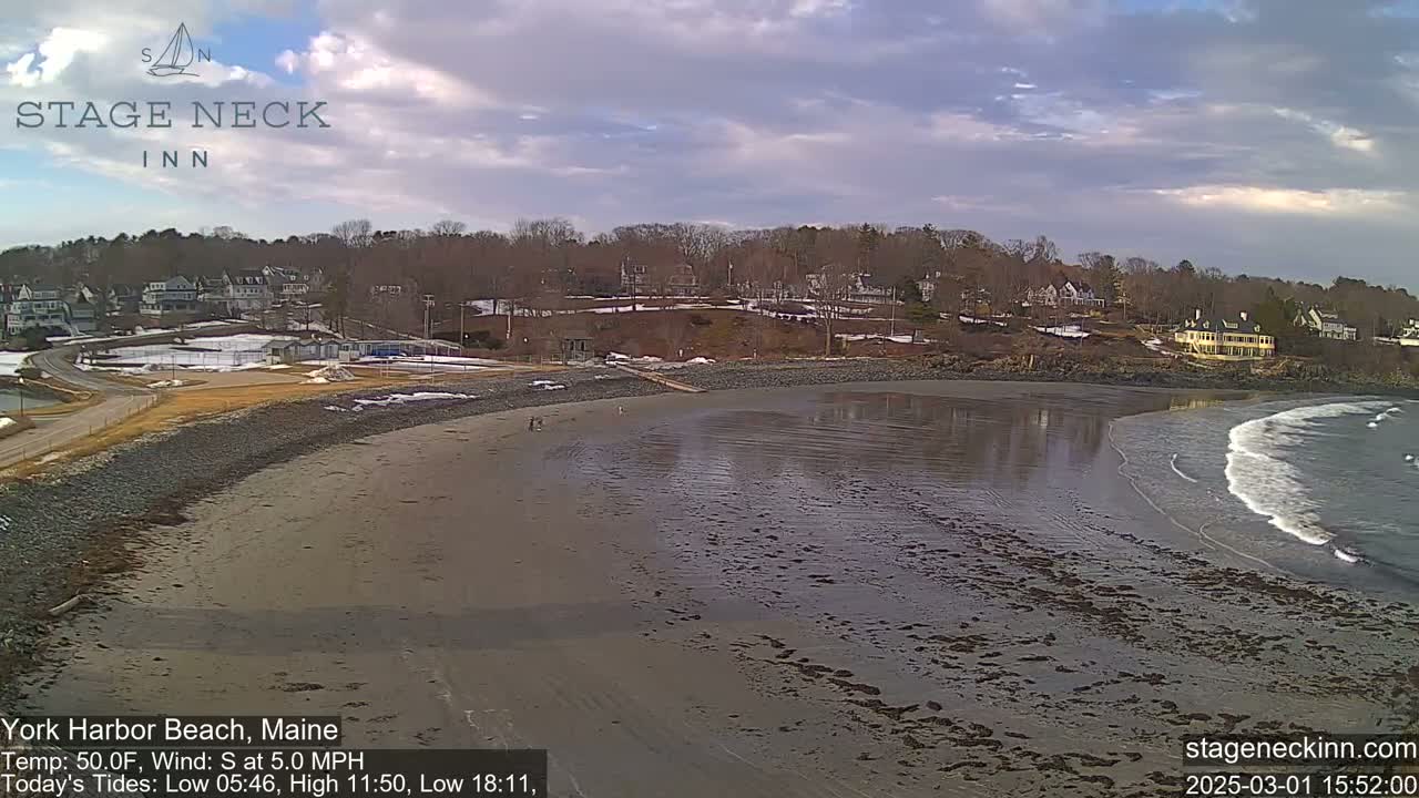 A mostly sandy beach with some exposed mudflats and small waves laps at the shore under a partly cloudy sky, with houses visible on a hill in the background.