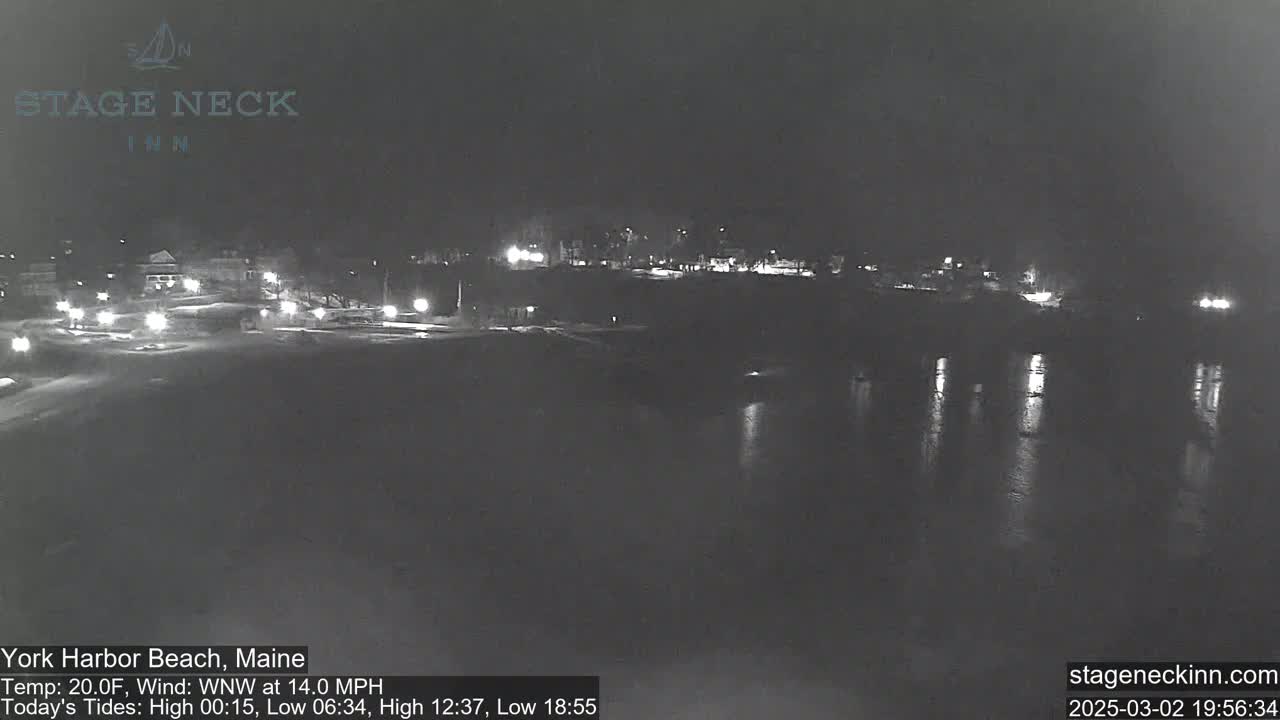 A nighttime view of a dark, calm body of water reflecting faint lights from a distant shoreline, with a light wind blowing.