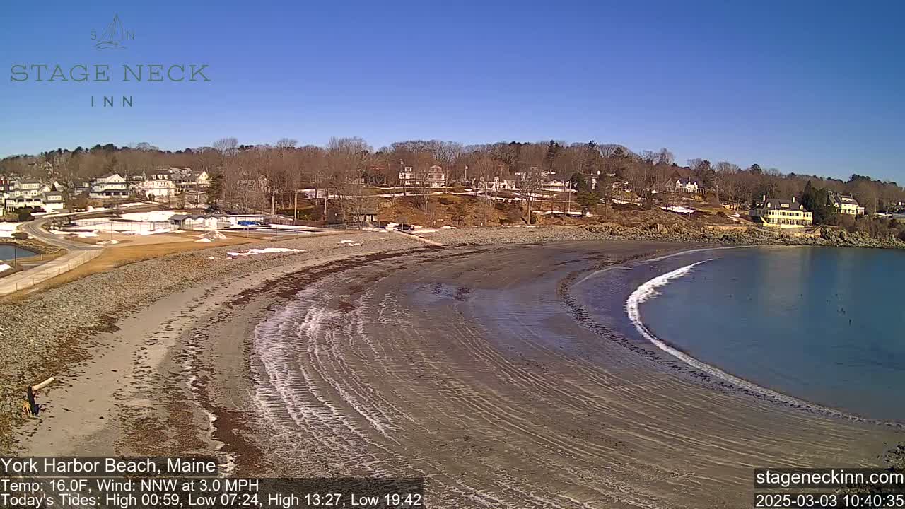 A sandy beach curves into a calm bay under a clear blue sky, with a small town visible in the background.